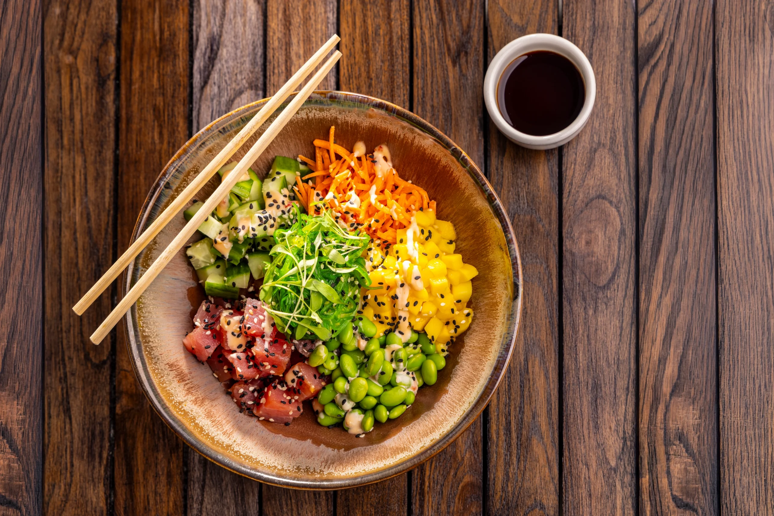 An ahi tuna poke bowl with cucumber, shredded carrot, mango, edamame and seaweed salad, served with chopsticks and soy sauce